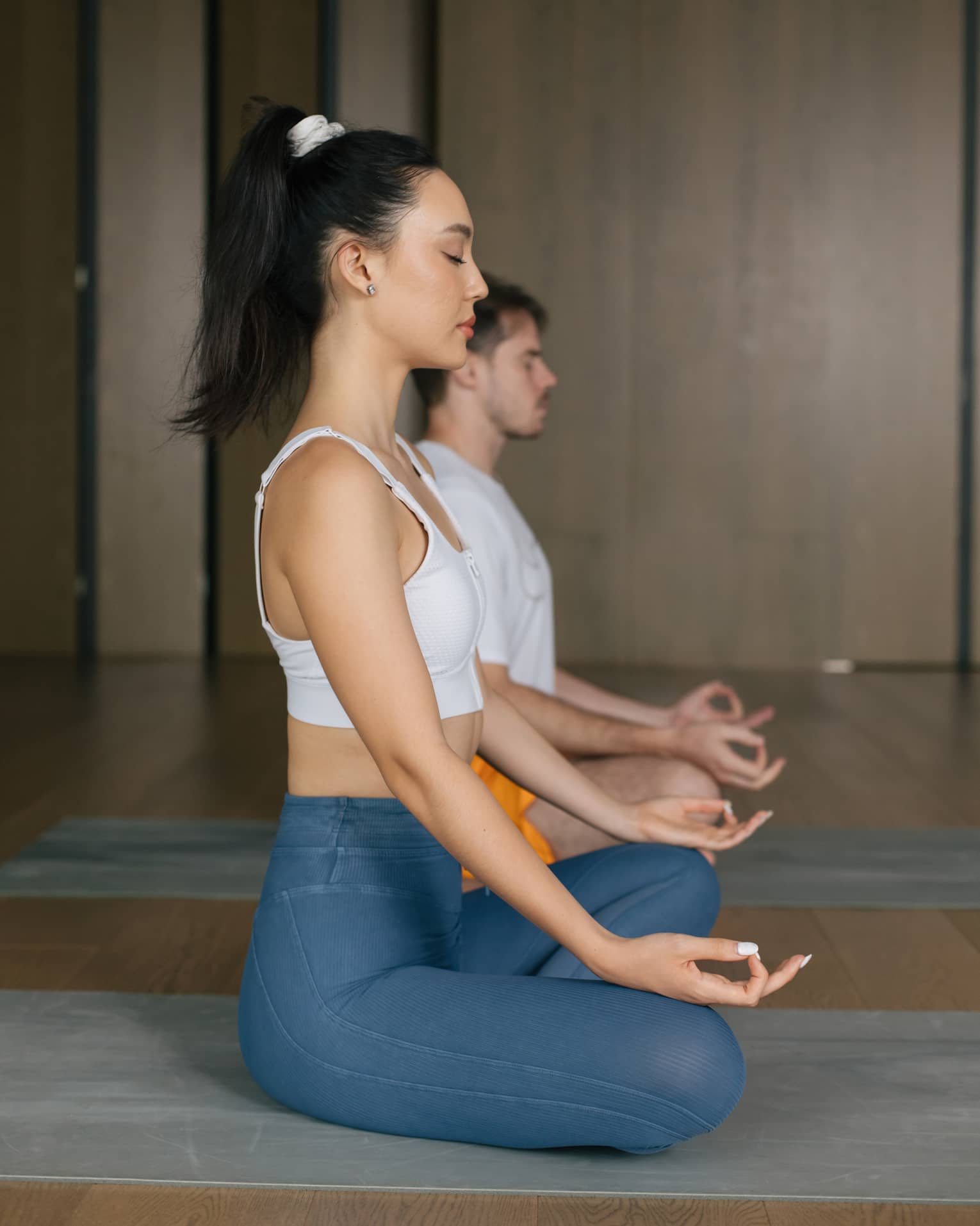 Woman with dark hair tied up in a ponytail and wearing a white tank top and blue yoga pants sits cross-legged on a yoga mat with her hands on her knees and eyes closed, a man wearing a white t-shirt and orange shorts does the same behind her