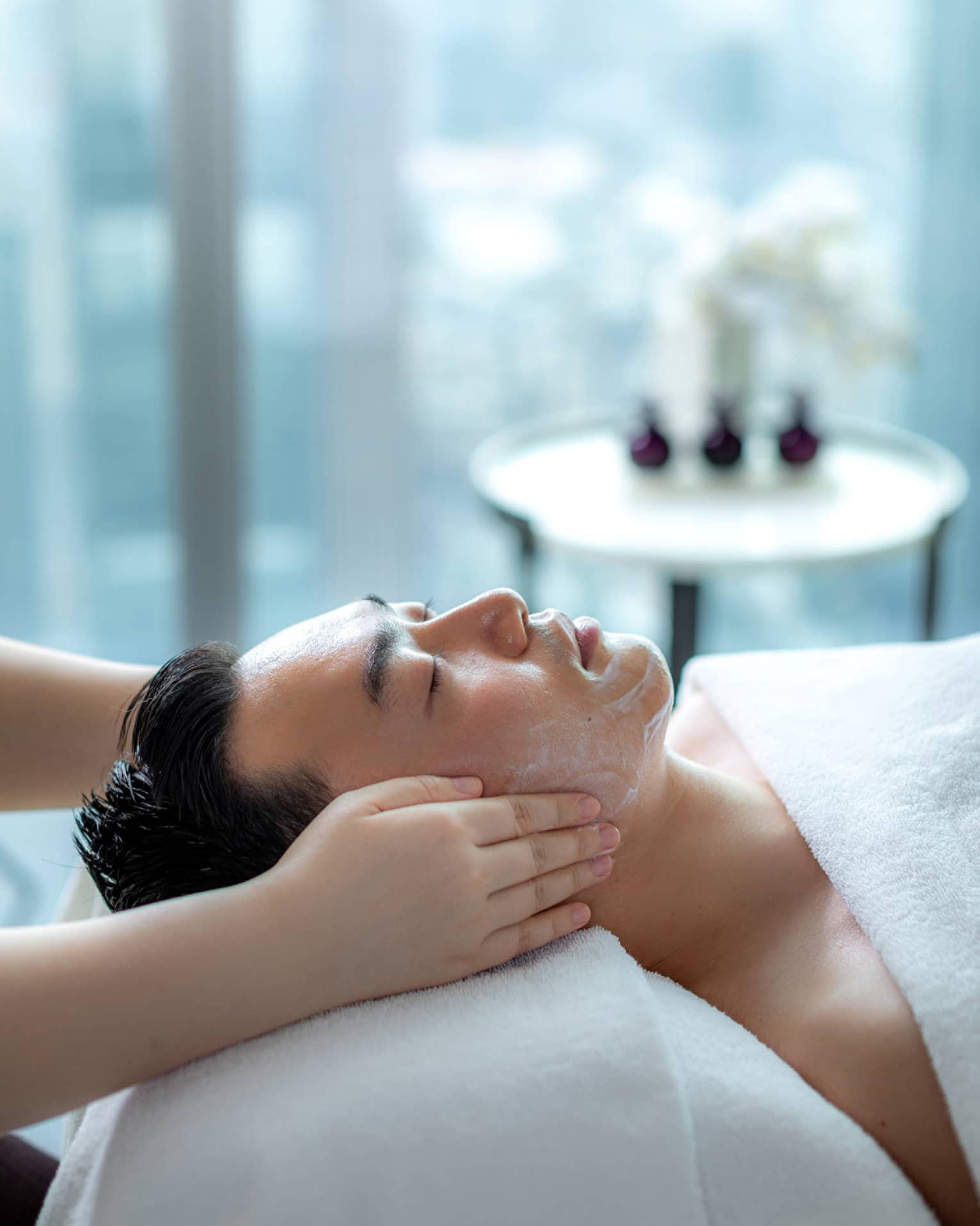 Hands rest on man's cheeks as he lays on massage table by glass window with daytime city view 