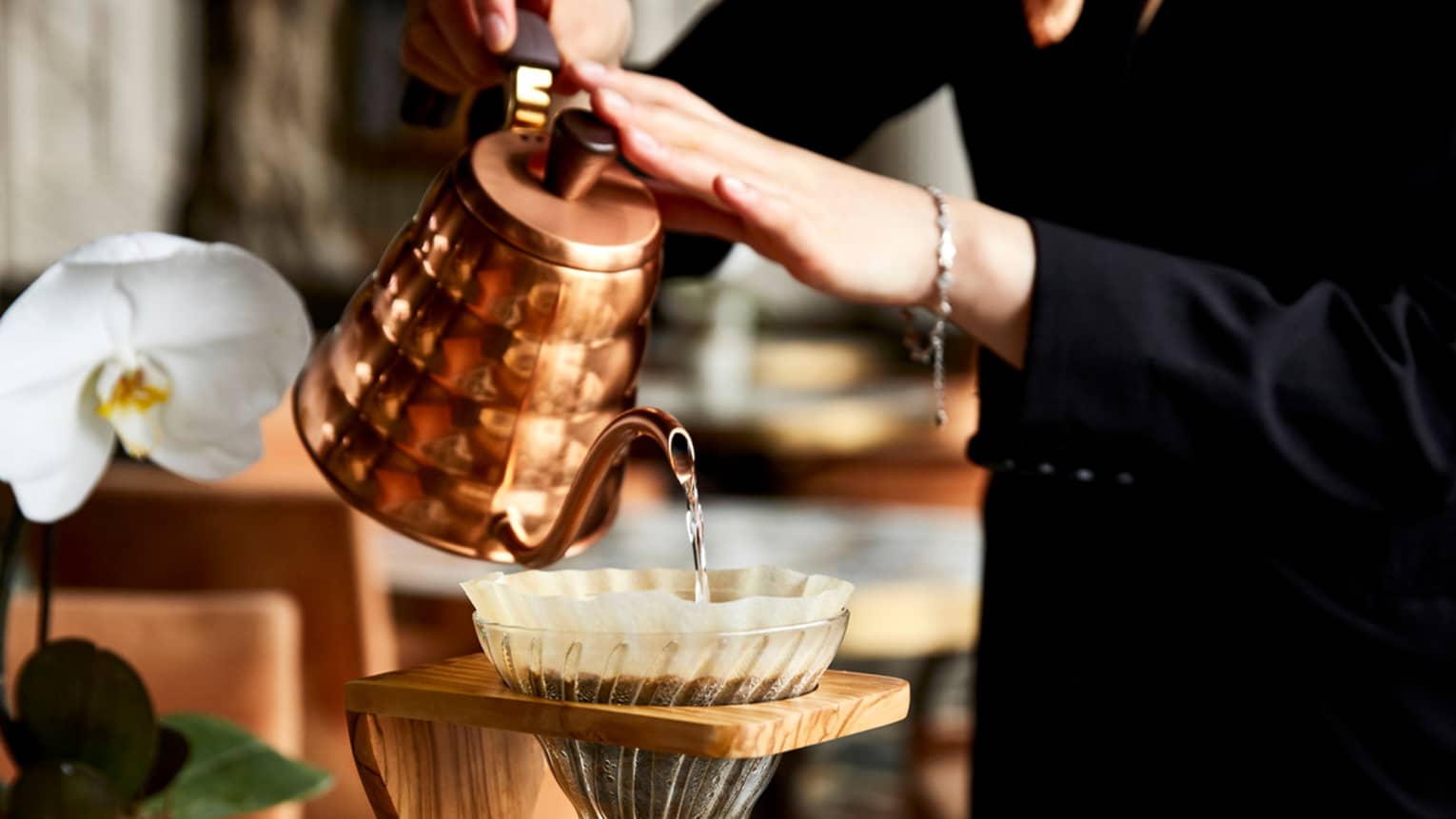 A woman pours water into a filter filled with coffee grounds, which drips into a glass container of hot coffee