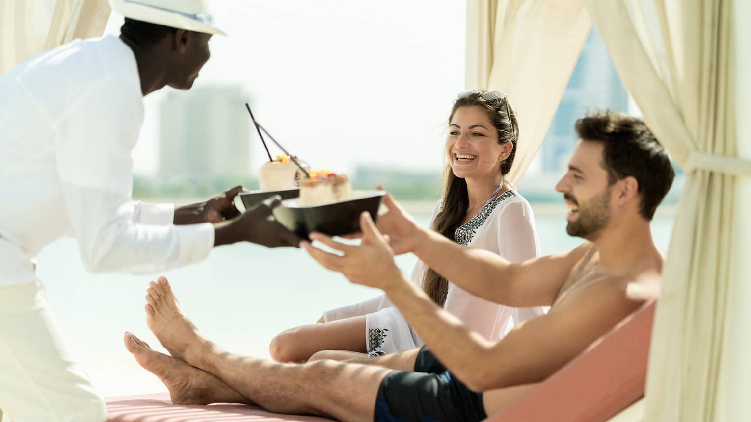 A four seasons staff hands two dishes to a man and woman as they lounge poolside