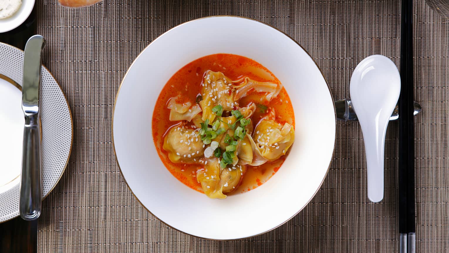 In-room dining white bowl with dumplings in red sauce, sprinkled with white sesame seeds, green onions