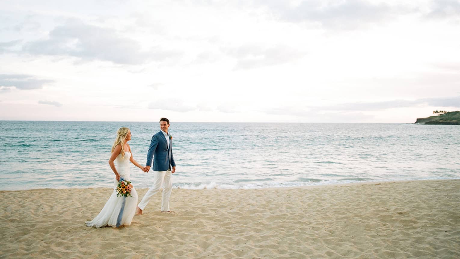 A groom is holding a brides hand as they walk along a beach.