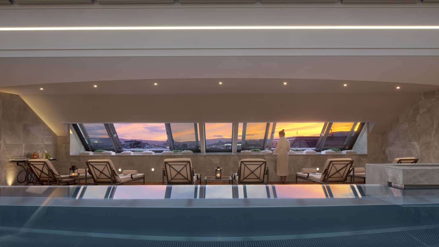 Woman in robe stands on deck beside blue indoor swimming pool, lounge chairs, looking at sunset through window
