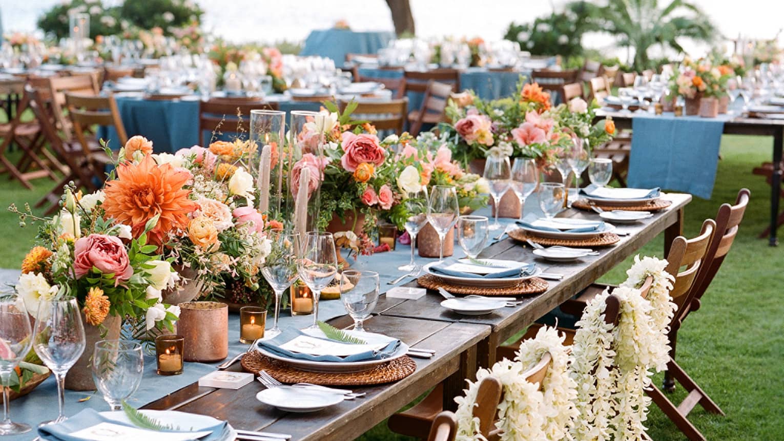 Close-up of outdoor banquet dining table with flower arrangements, white floral leis draped over chairs
