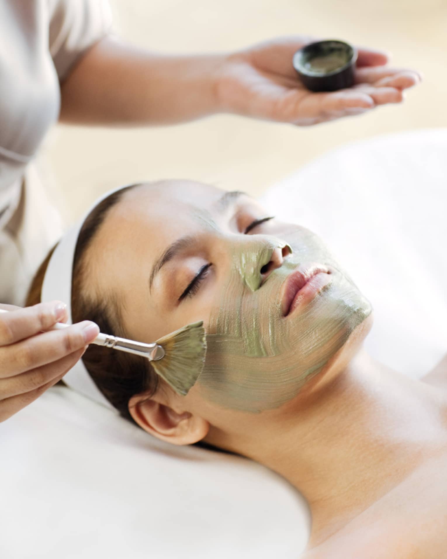 Spa staff holding brush paints green mask on woman's cheeks, chin, nose 