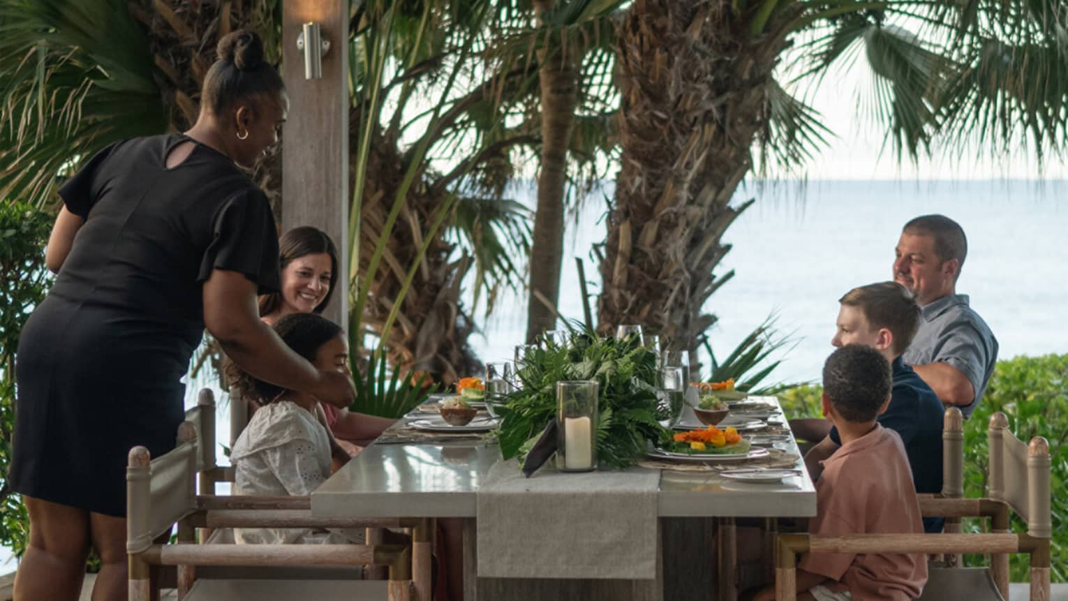 A family smiles while being served a meal at an outdoor dining table with an ocean view