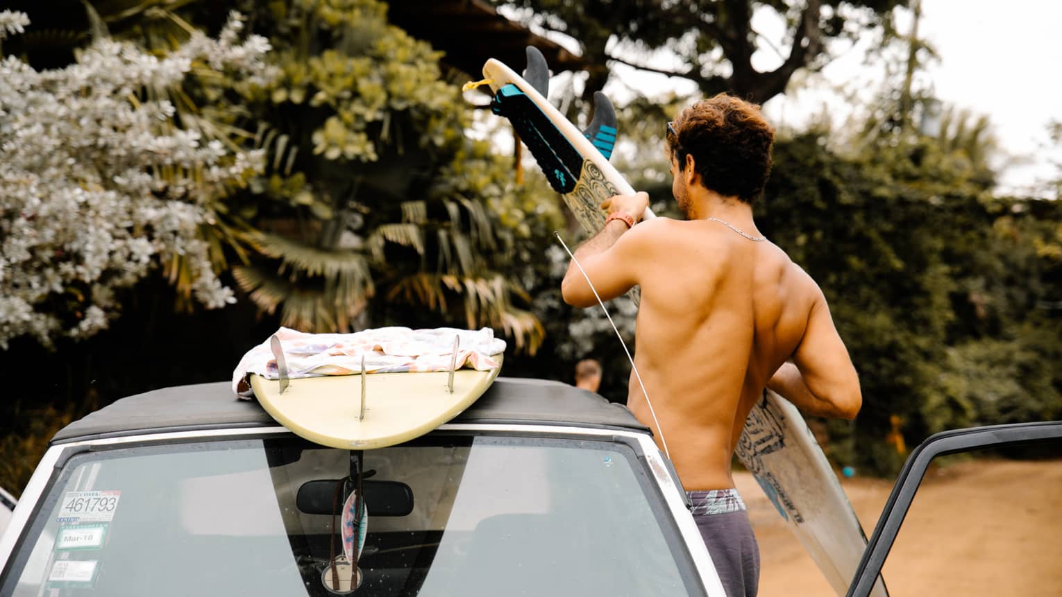 Opposite green and white foliage, a beachwear-clad surfer grips a surfboard, preparing to load it onto the roof of a car. 