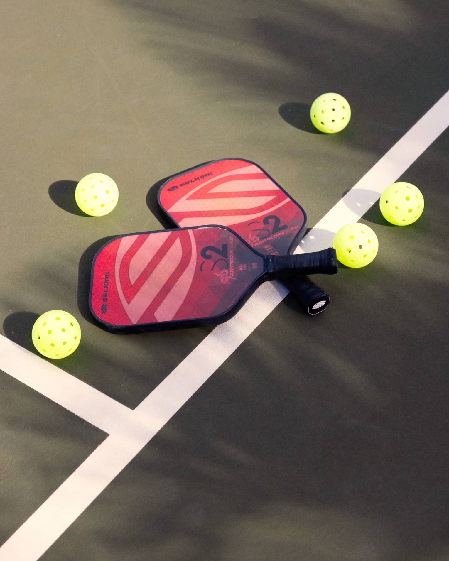 Close-up of two red and pink pickleball racquets, handles crisscrossed on a white court line, surrounded by five pickleballs.