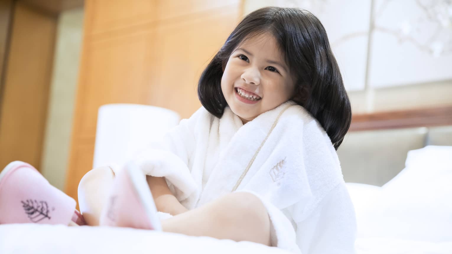 Smiling little girl wearing white bath robe and pink slippers sits on bed