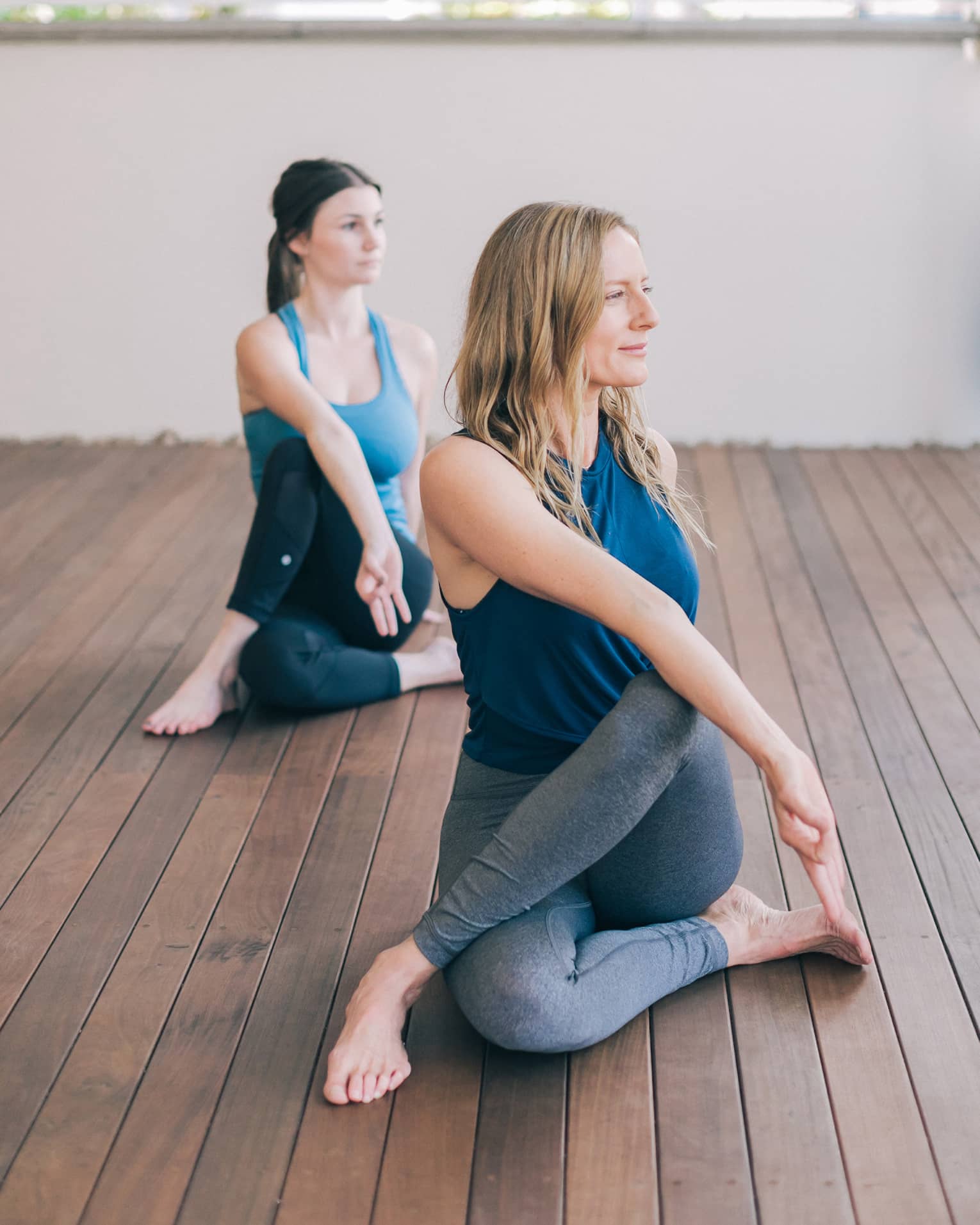 One blonde woman and one brunette woman sit with one leg crossed over the other with their bodies turned to one side and their front hands forming a lam mudra