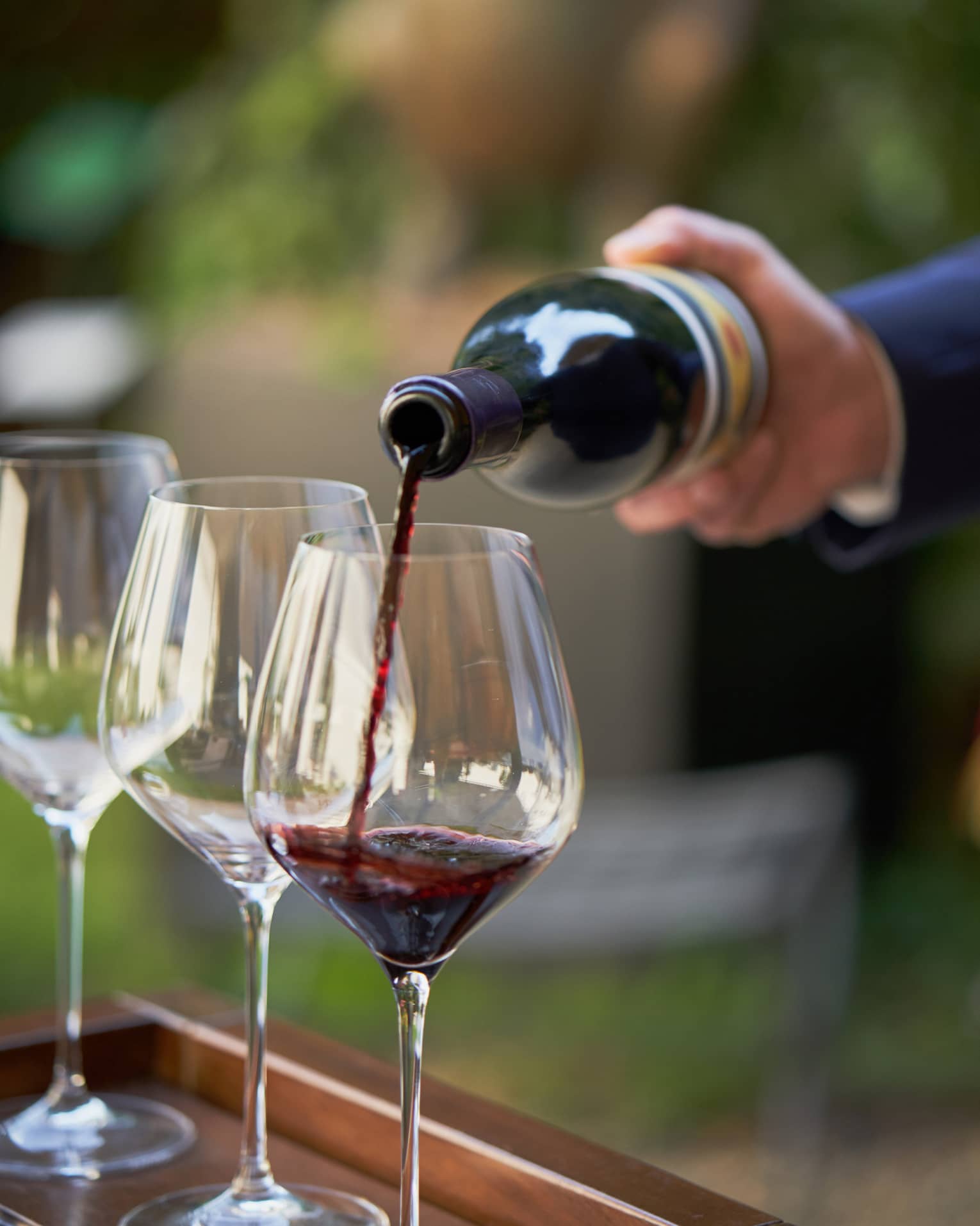 A close up of hands pouring a bottle of red wine into a clear wine glass that rests on a wooden tray, outdoors