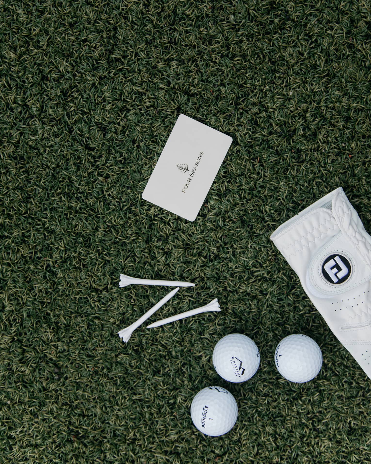 On green turf, a Four Seasons card, three golf balls, three tees and a white leather golf glove dotted with tiny holes.