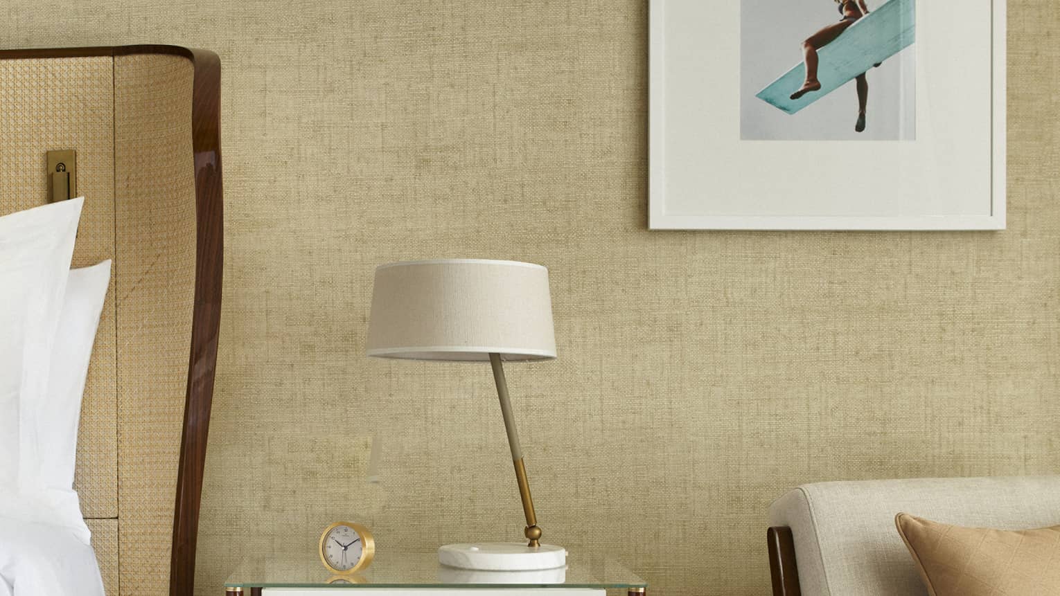 Next to a bed sits a nightstand with lamp, arm chair, square framed artwork on wall