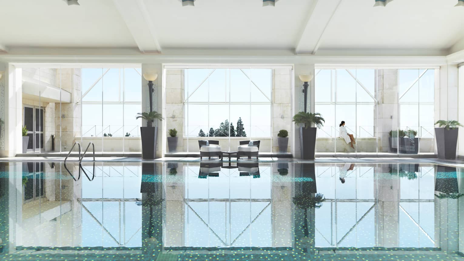 Indoor swimming pool reflecting three tall windows, woman in white bathrobe sits on ledge