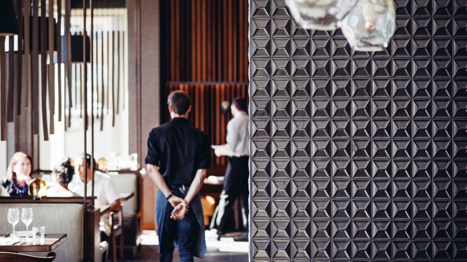Server walks with hands behind back past dining tables, beyond decorative metal wall