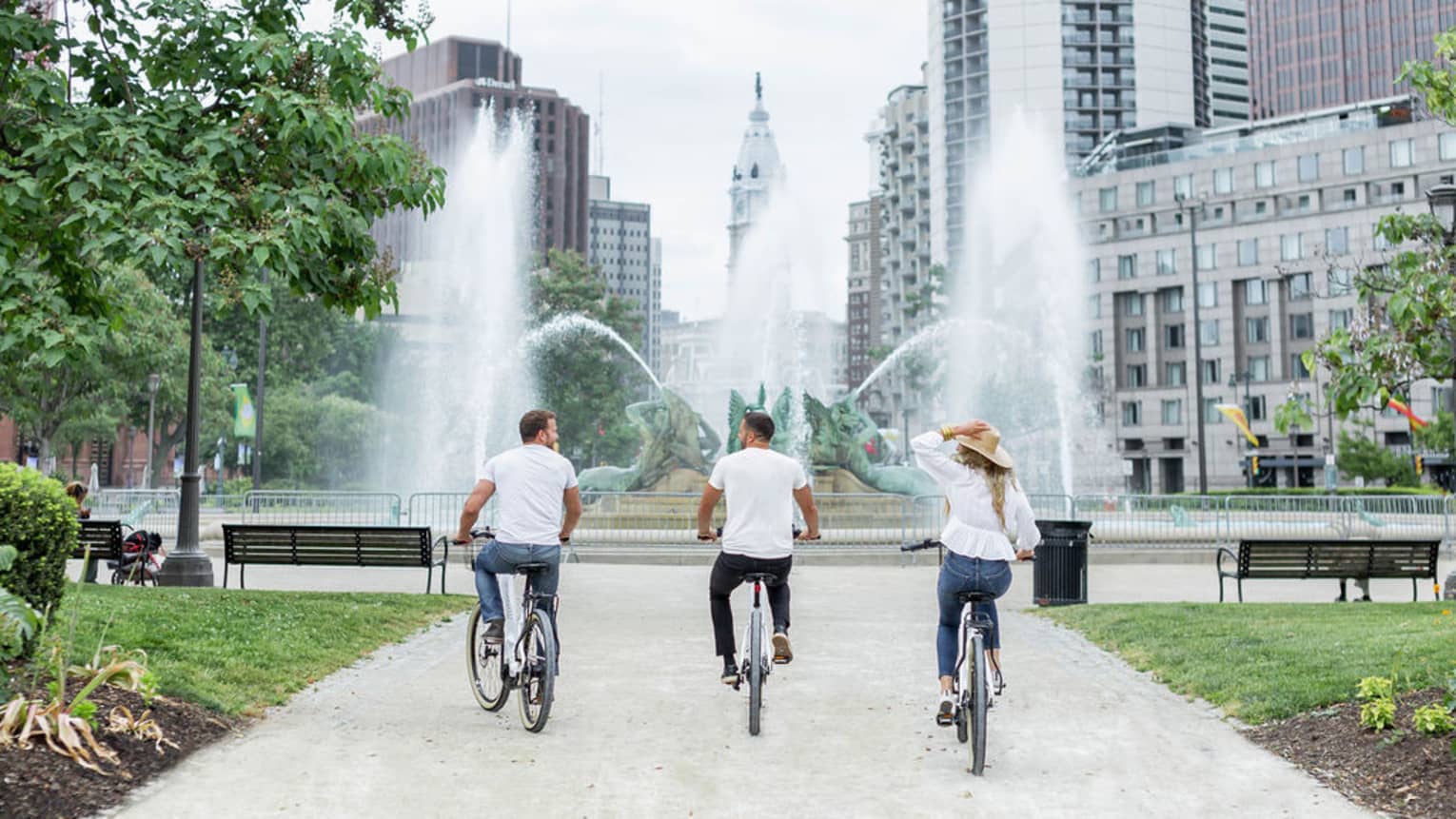 Three guests riding bikes towards a large fountain with the Philadelphia skyline in the distance