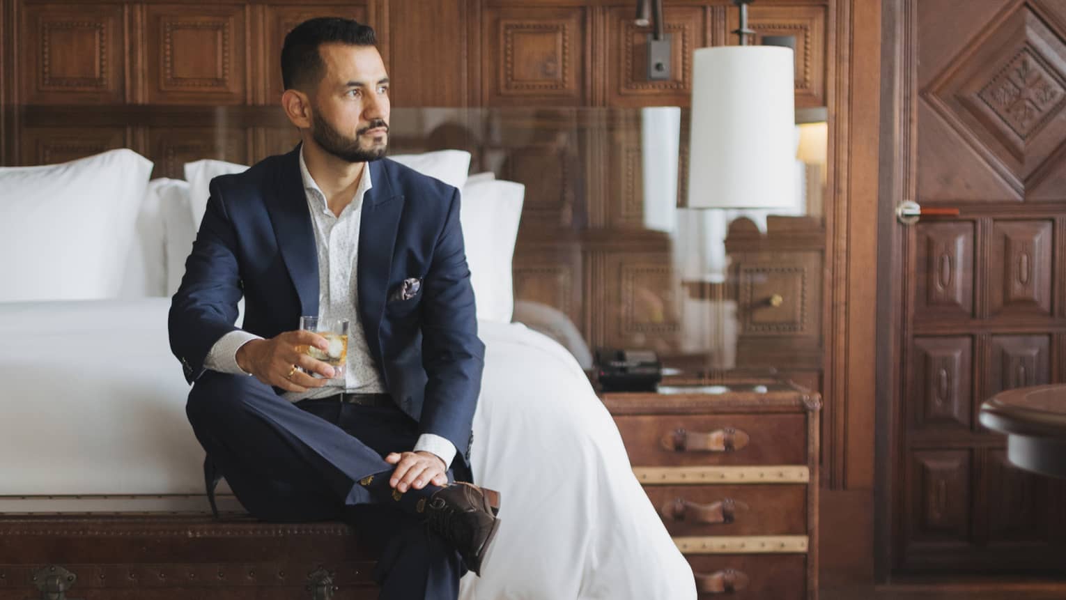 A bearded man wearing a navy blue suit with a white shirt sits ankle-on-knee and holding a drink by the bed in a wood-paneled guest suite