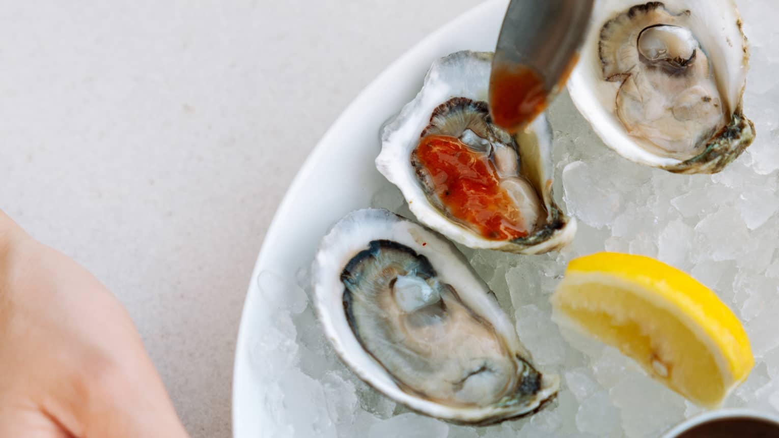 A manicured hand by a bowl of shucked oysters drizzled with red sauce, a lemon wedge and sauce cups on a bed of crushed ice.