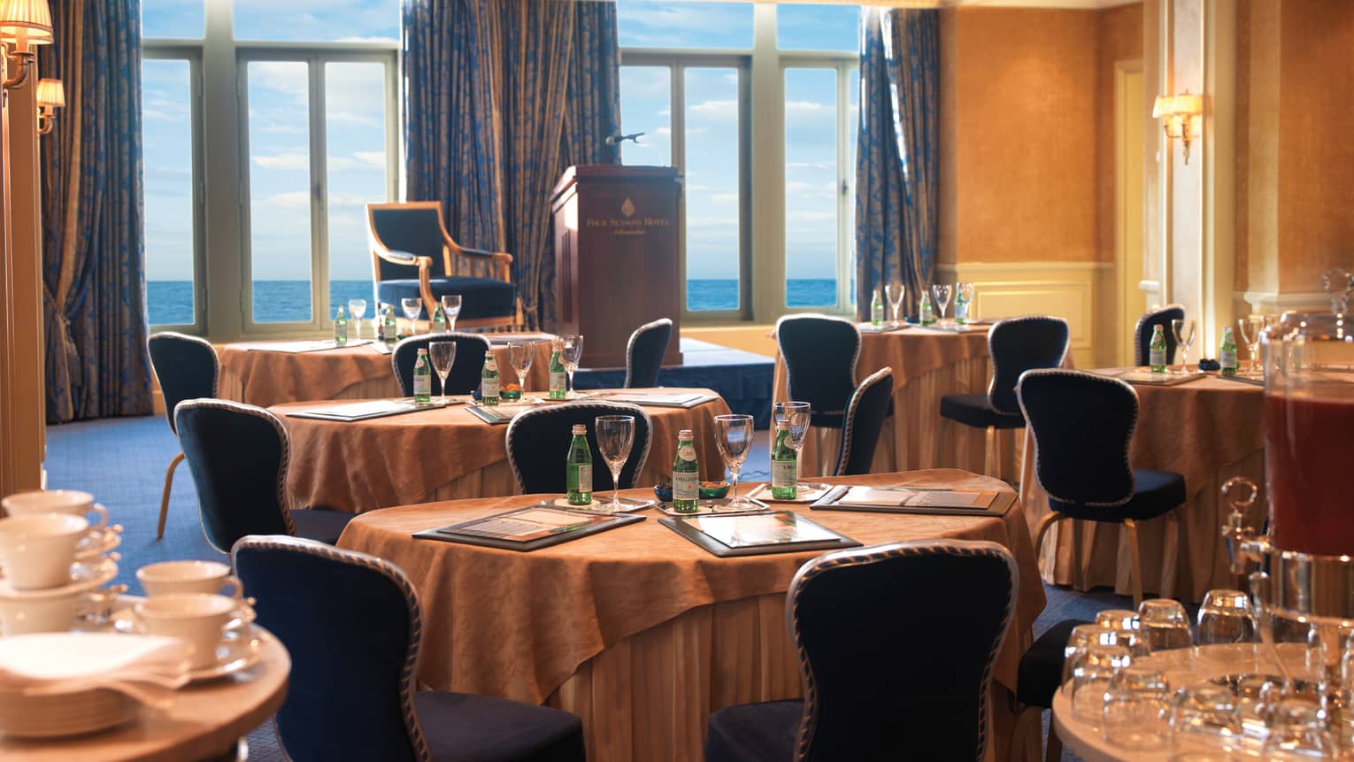 Sun-lit meeting room with round tables, royal blue chairs, podium facing floor-to-ceiling window and ocean views 