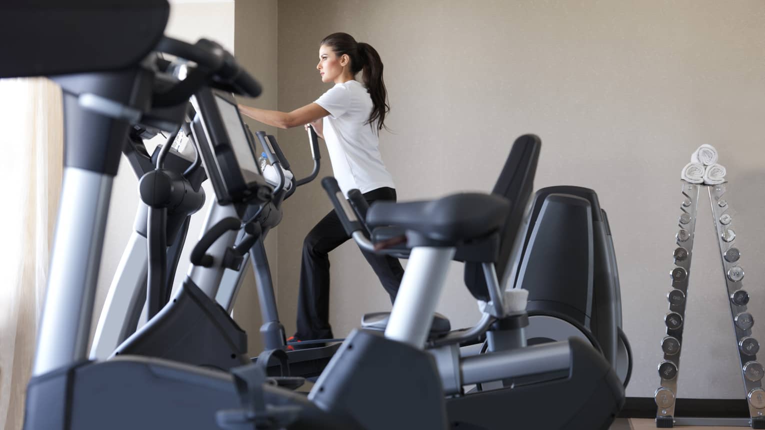 Woman in white shirt, black pants works out on cardio elliptical machine in sunny Fitness Centre 