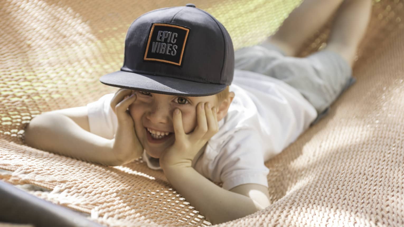 Child laying on hammock on stomach, face in hands and wearing a black cap