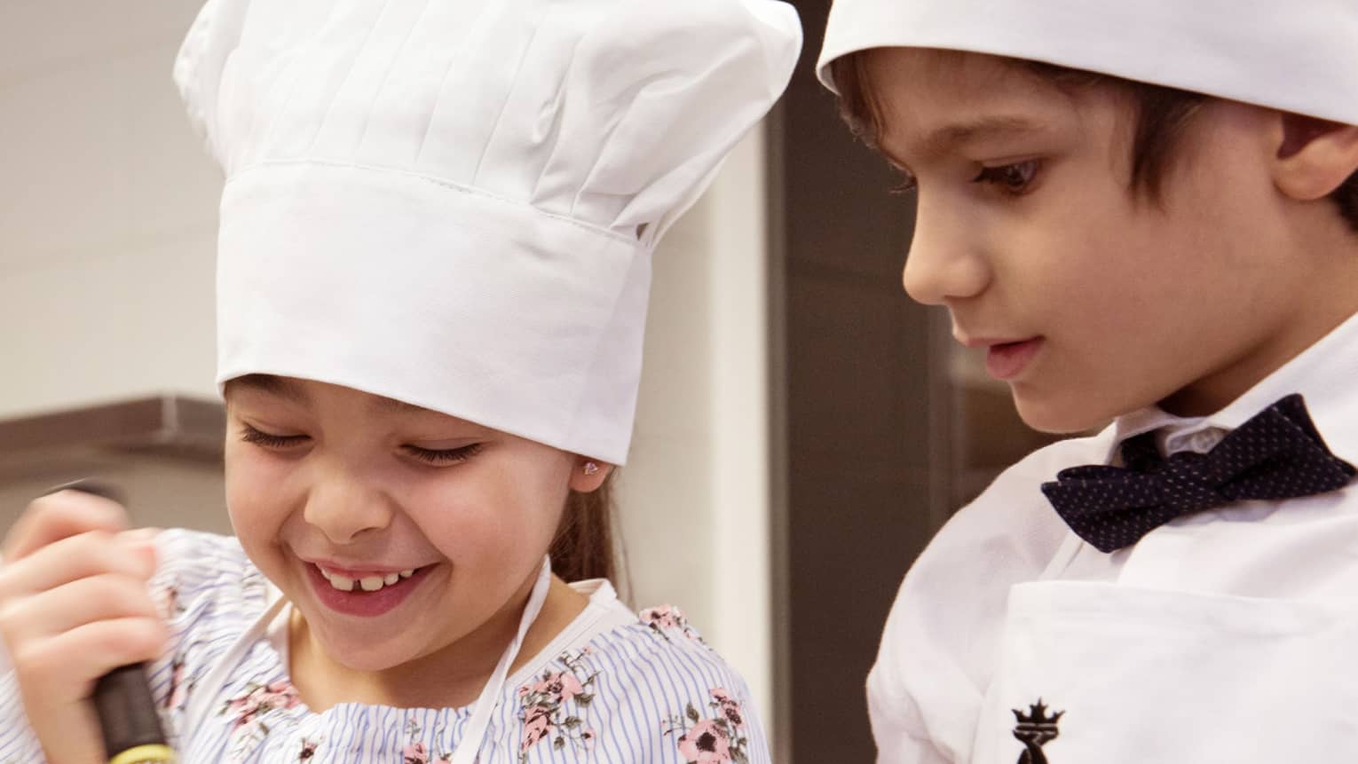 Girl and boy in chef hats; girl stirring with whisk, boy watching
