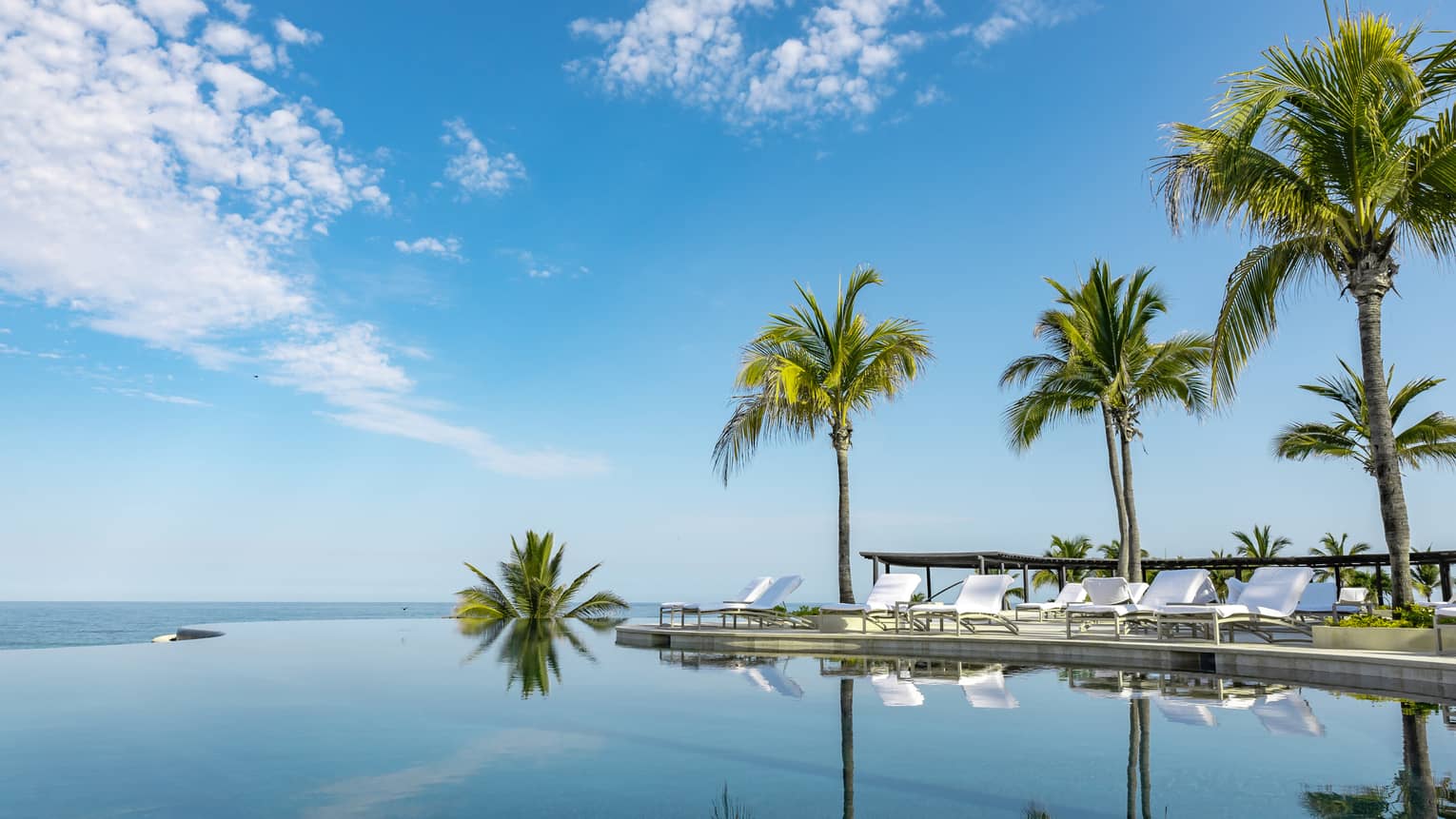 A calm infinity pool reflects the blue sky, clouds and palm trees