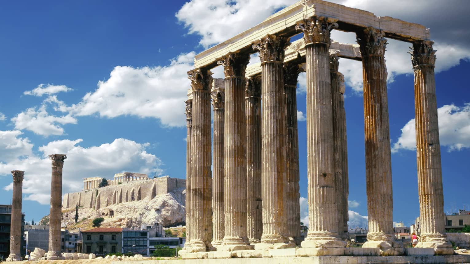 A temple with towering columns featuring intricate designs; more ruins and the Parthenon prominent in the background.