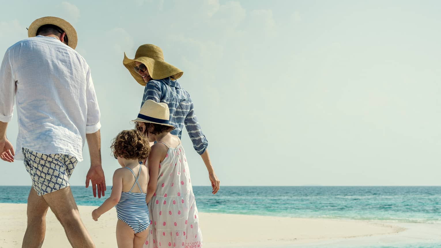 Family wearing swimsuits, sundresses and sun hats walk along white sand beach