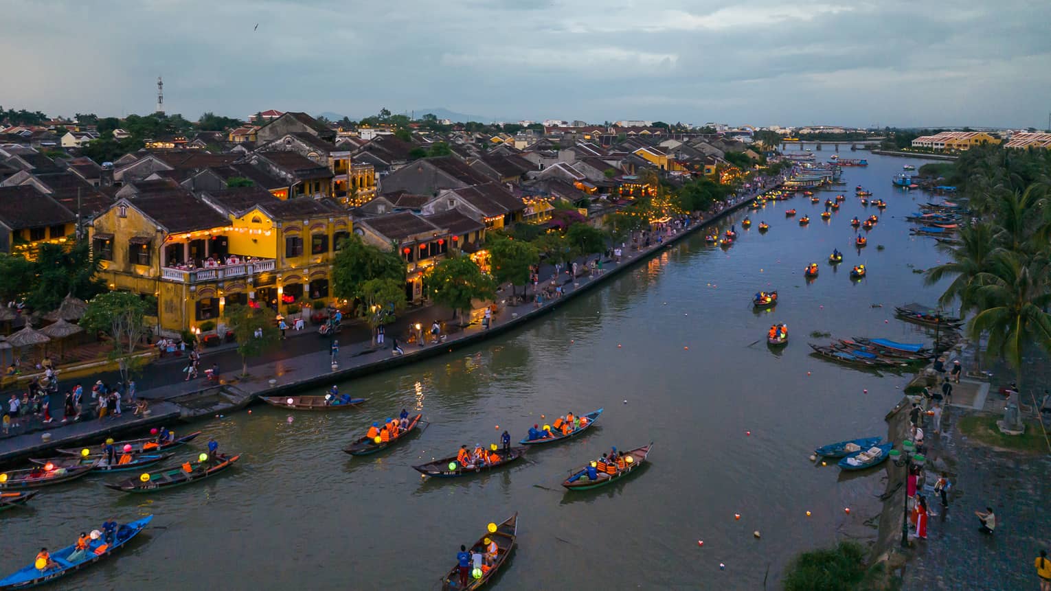 At dusk, lantern-lit canoes navigate past yellow buildings lit with red-and-yellow lanterns, along a bustling shoreline.