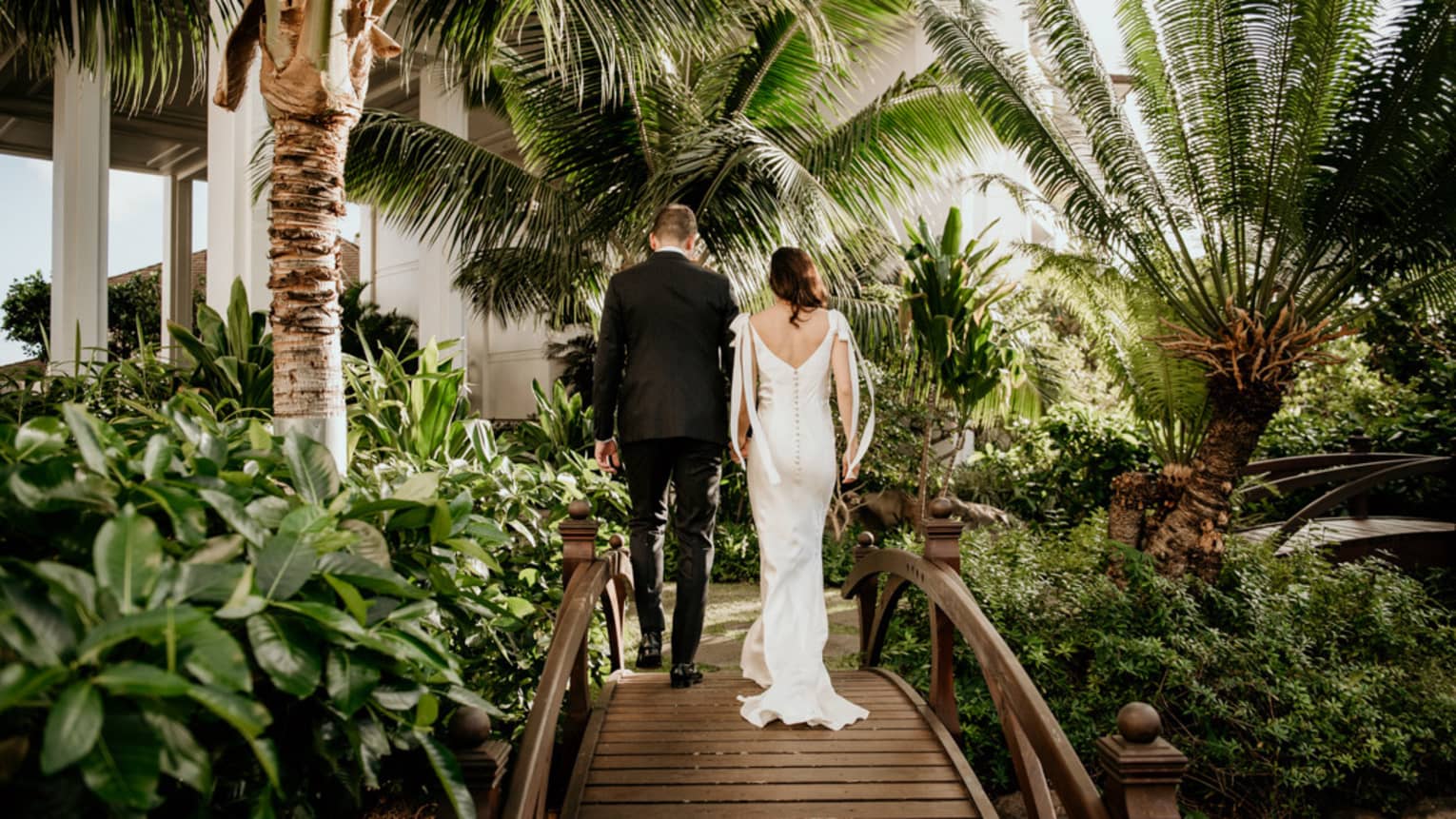 Bride and groom walk away from camera, over small wooden bridge, surrounded by tropical gardens