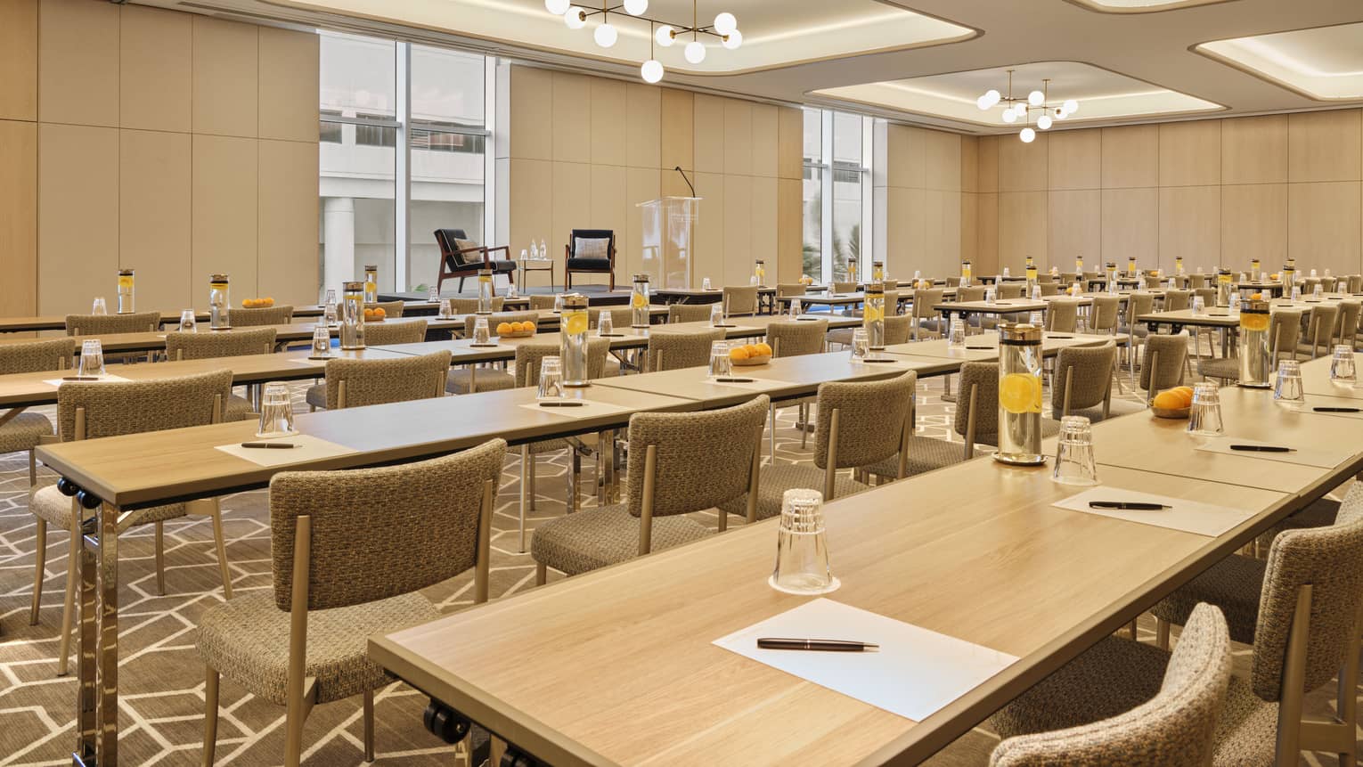 A large meeting room with long wooden tables, beige chairs and glasses of water in front of each chair.