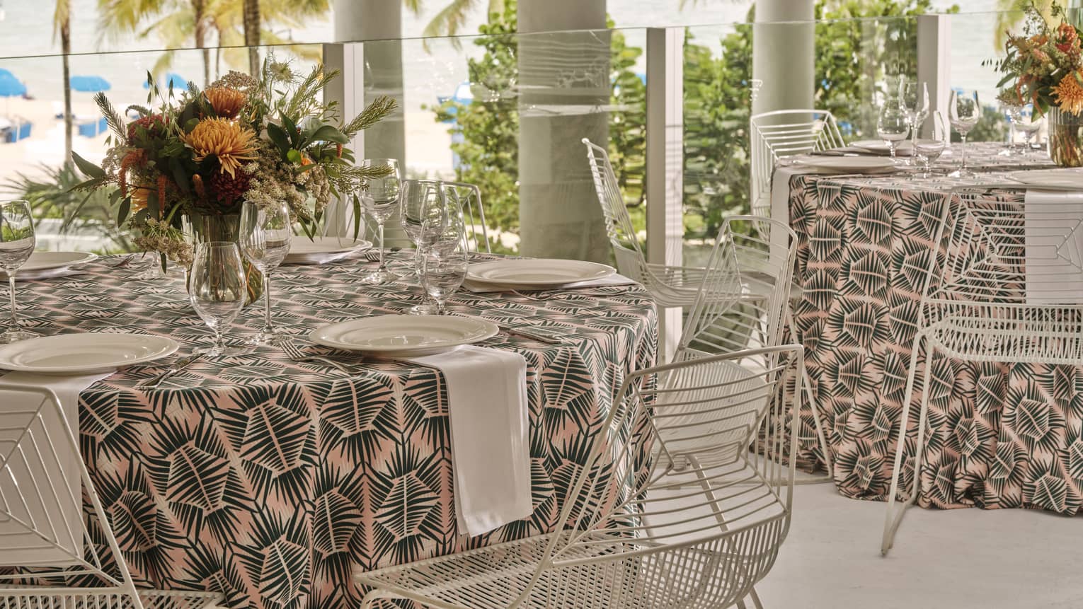 Round tables with table settings outside near a beach with palm trees.