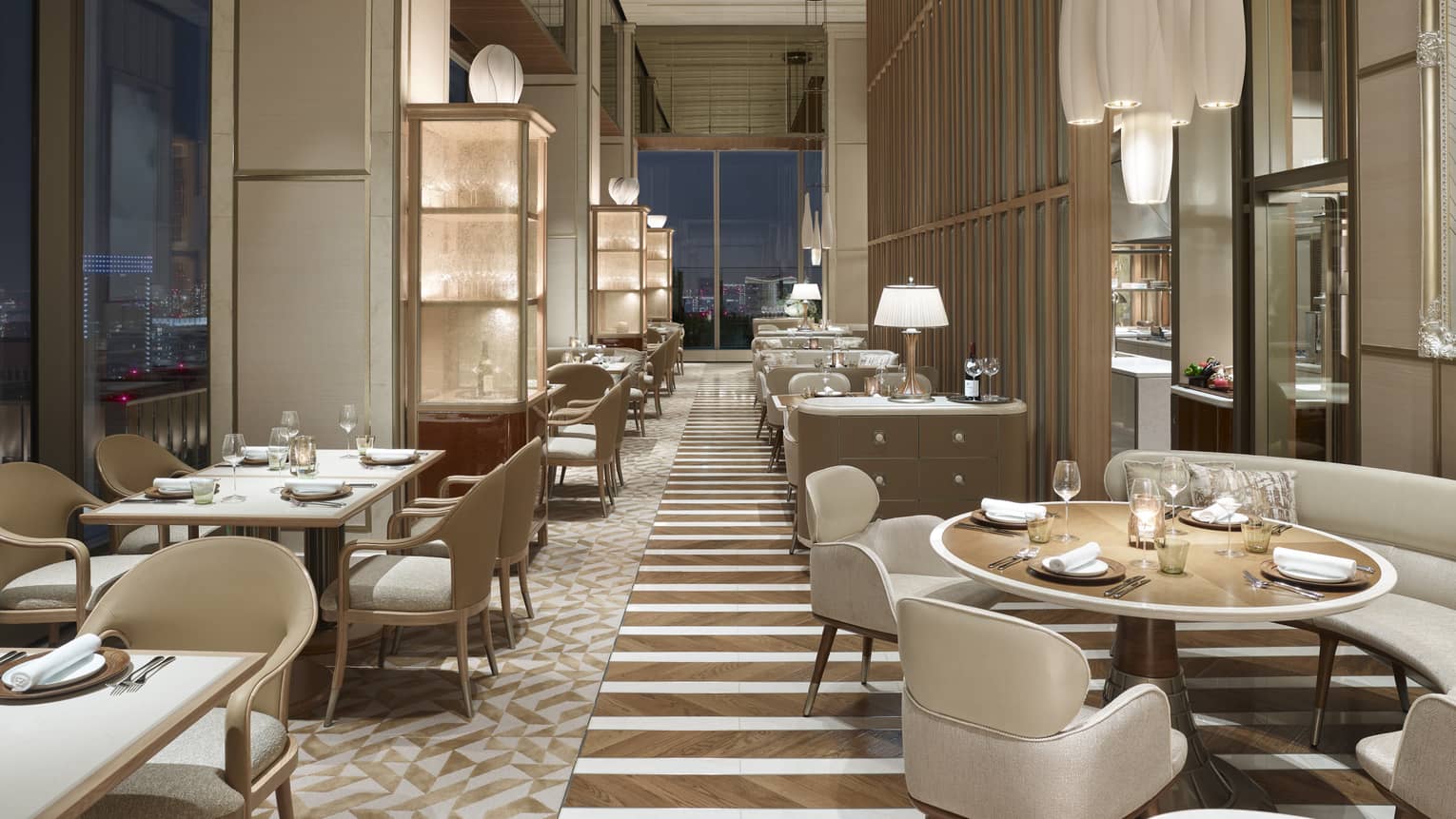 Restaurant with off-white chairs and sofas, elegant chandeliers, windows with nighttime views