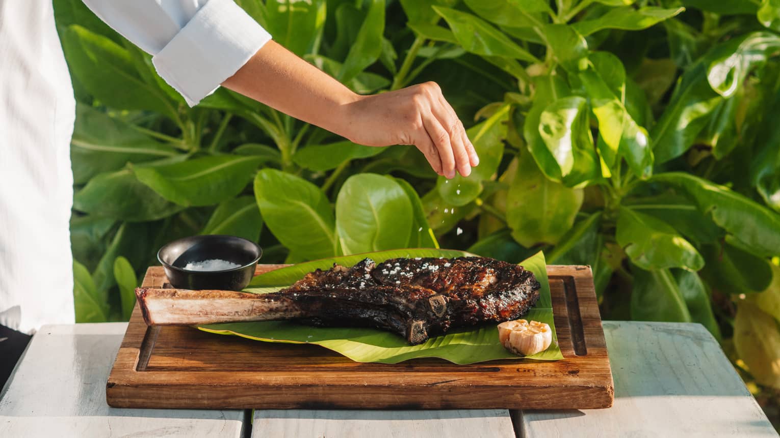 Chef sprinkling seasoning on a grilled tomahawk rib-eye steak on banana leaf