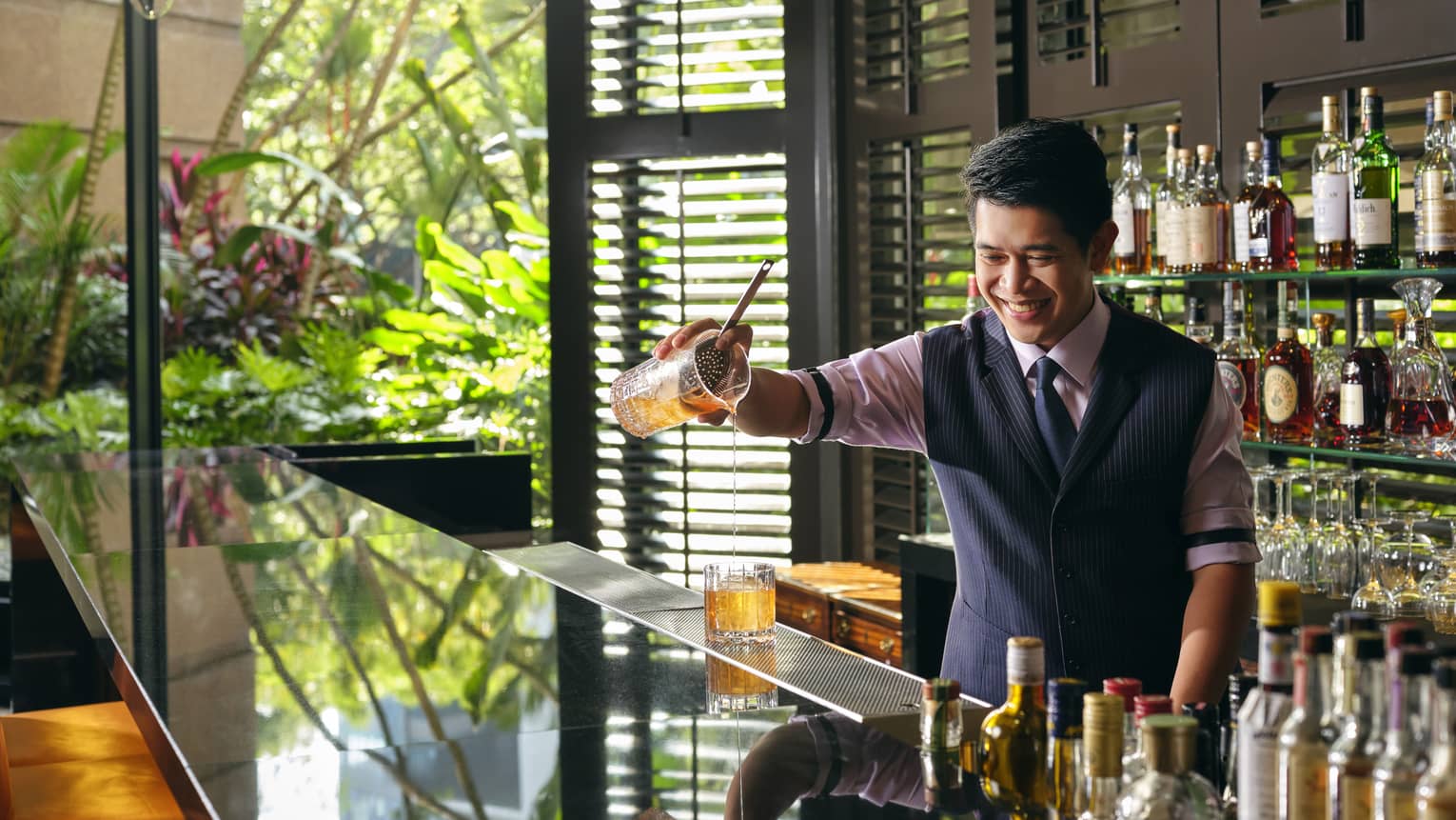 A smiling bartender pours a Sidecar cocktail, a display of liquor bottles behind him, light streaming in from the windows to his right 