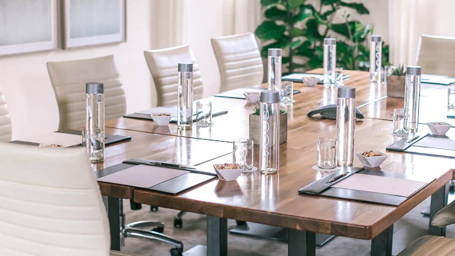 A rectangular boardroom table set with clear glass water bottles and black folders in a naturally-lit room