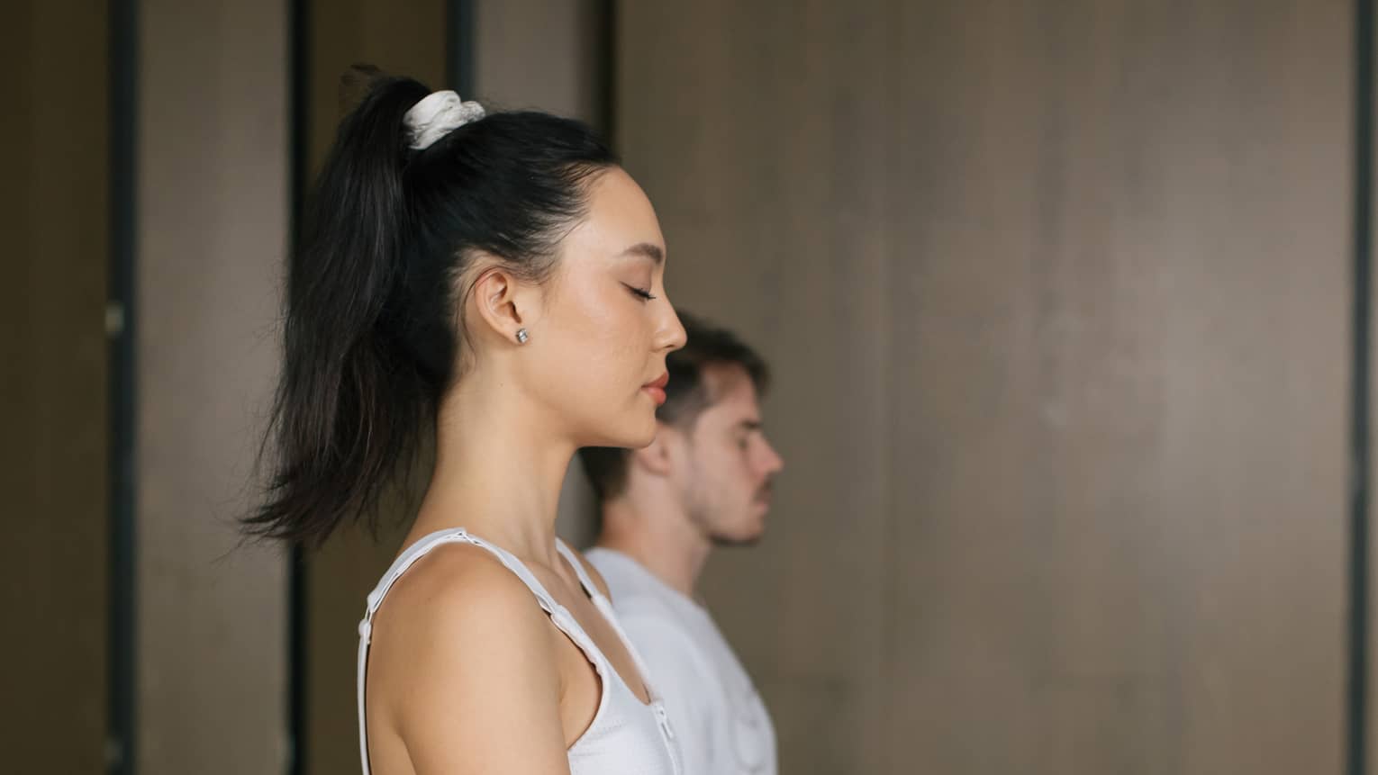 Woman with dark hair tied up in a ponytail and wearing a white tank top and blue yoga pants sits cross-legged on a yoga mat with her hands on her knees and eyes closed, a man wearing a white t-shirt and orange shorts does the same behind her