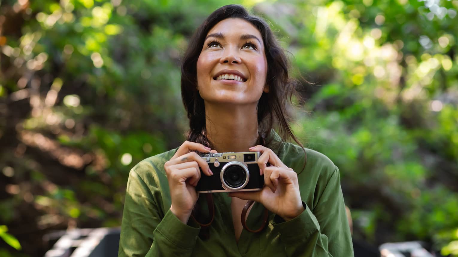 Woman with long dark hair wearing a green button-down shirt holds a camera as she looks upward in the forest