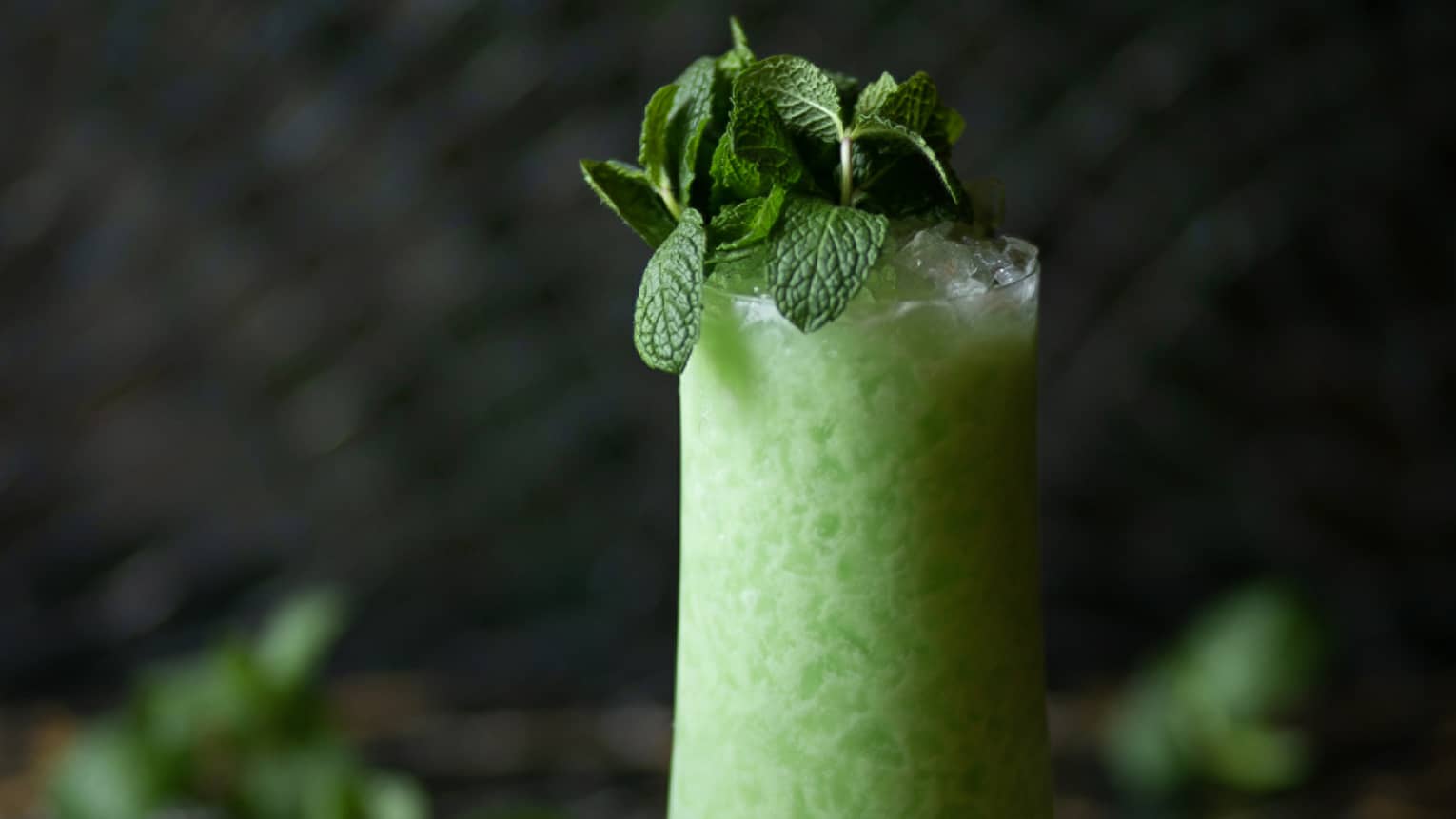 A neon green icy cocktail in a tall glass garnished with fresh mint leaves set against a dramatic dark background.