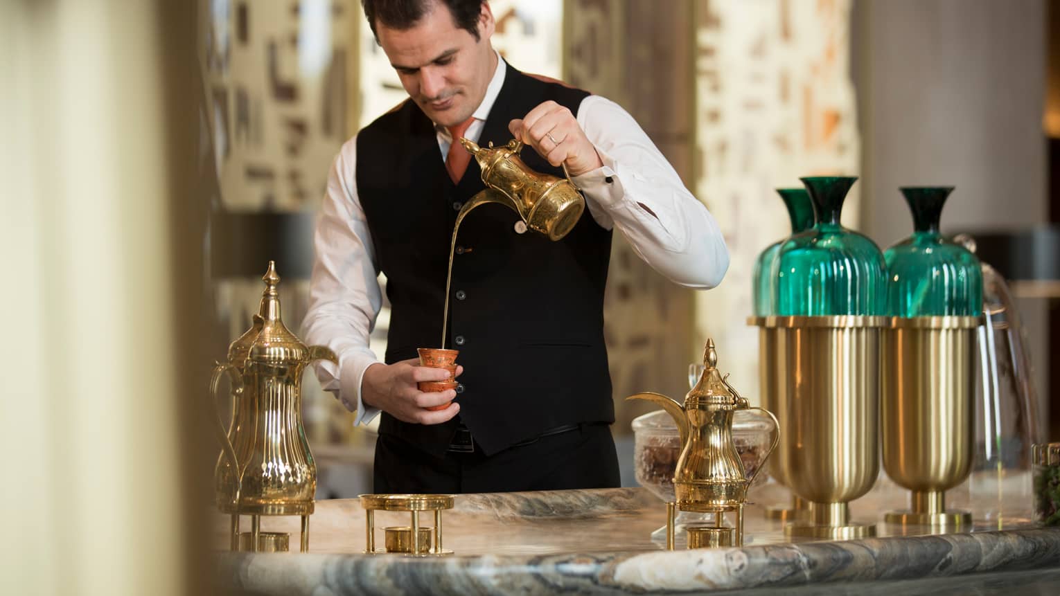Hotel staff in vest, tie pours tea from brass pot into small cup