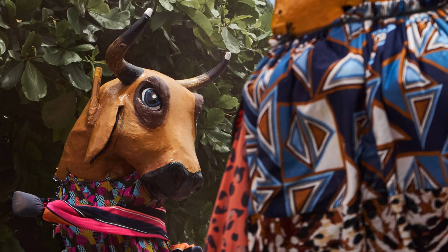 Person stands next to a tree wearing brightly colored patterned clothing and a large hand-made bull mask