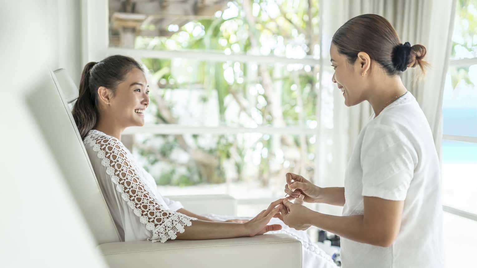 Woman sitting in white spa chait smiles to woman painting her nails during manicure