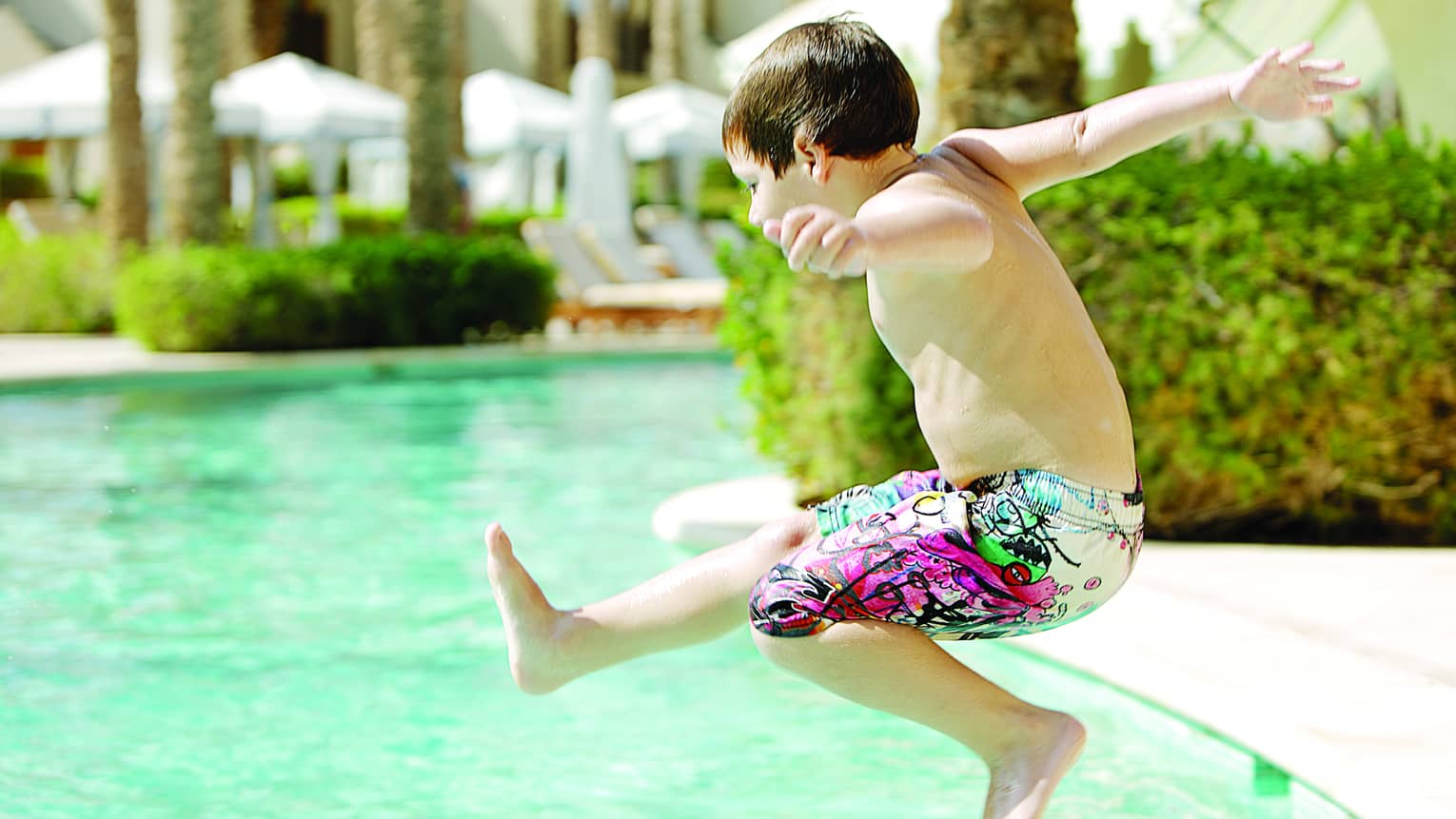 Boy wearing swim shorts jumps into sunny outdoor swimming pool