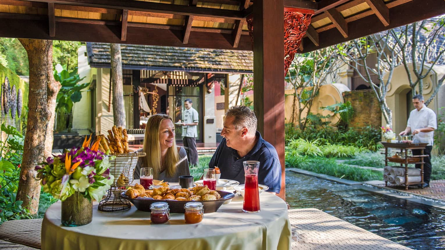 Smiling man and woman dine at table with baskets of pastries, breadsticks under wood gazebo