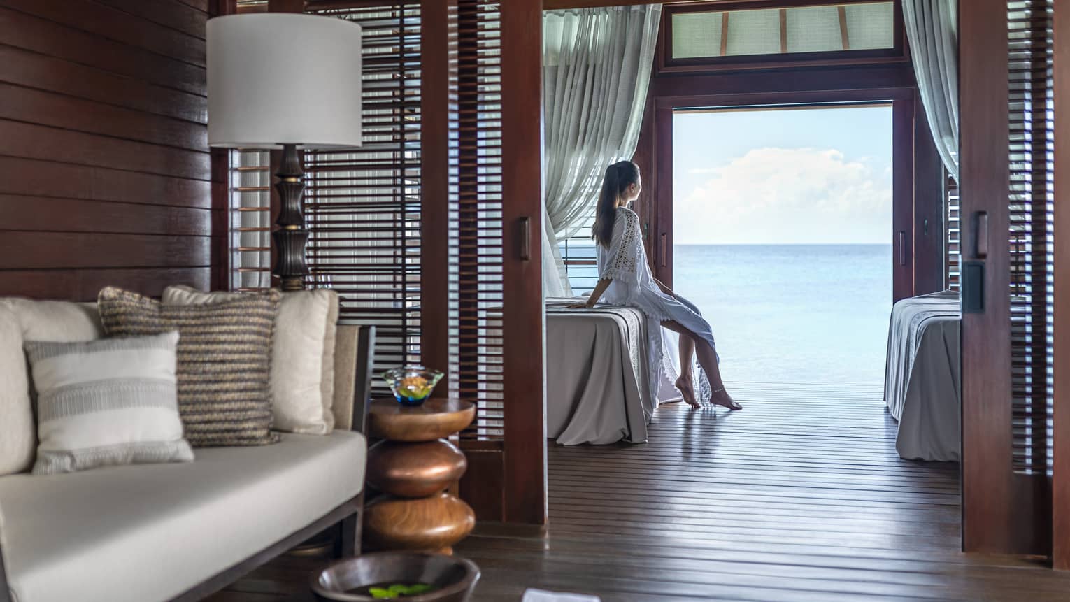 A woman leans on a massage bed on the far side of a spa treatment room and looks out through a large doorway onto the vast blue ocean