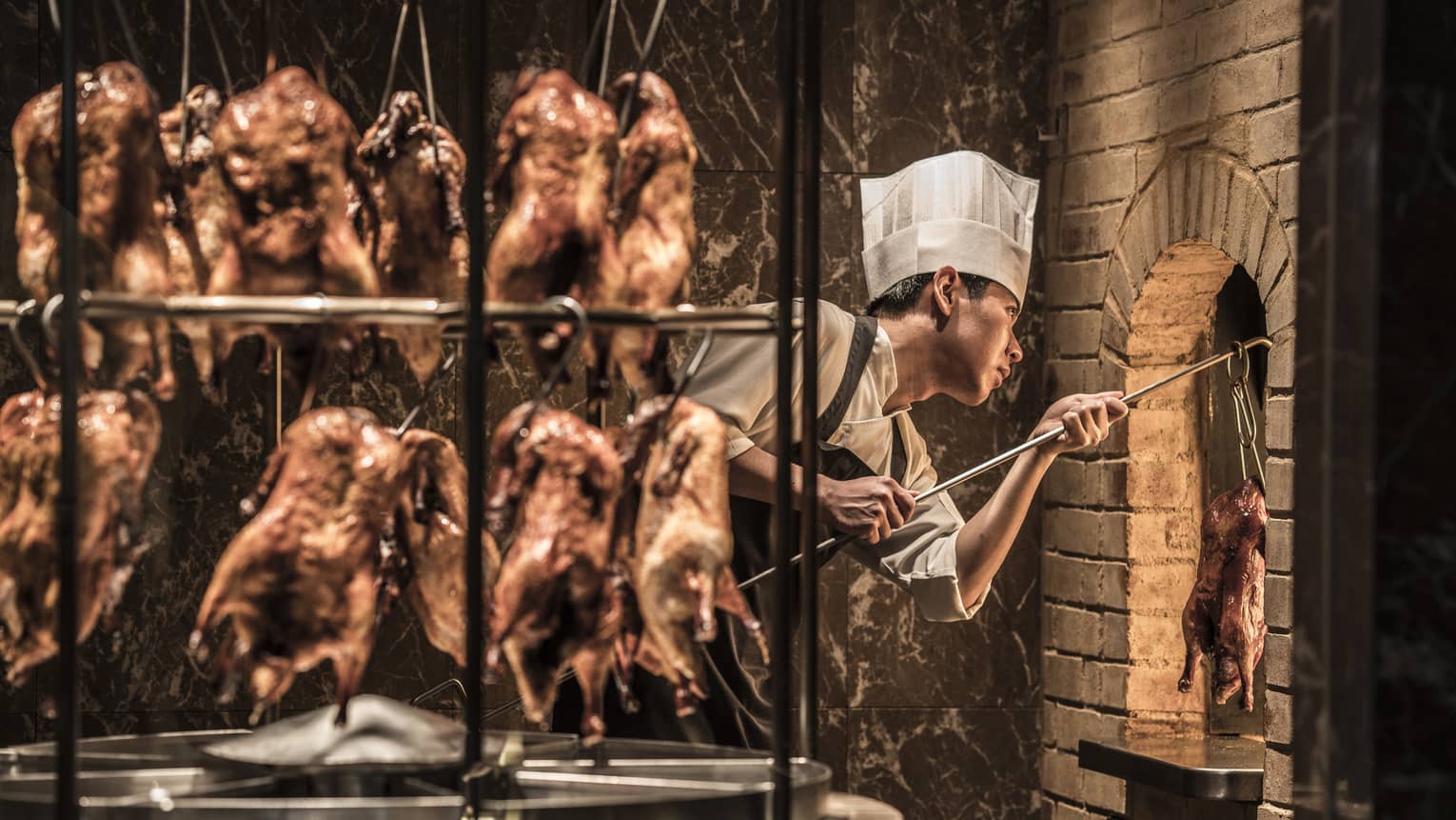 Rotisserie with hanging whole ducks in front of chef wearing white hat, taking duck from brick oven