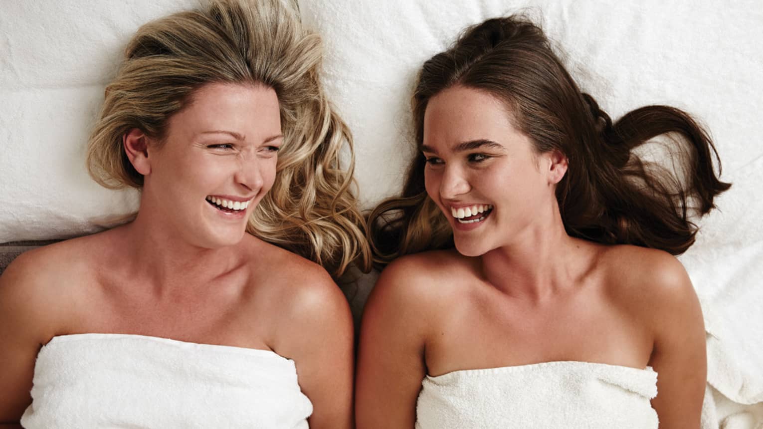 Two laughing women wearing white towels lie on massage table in spa