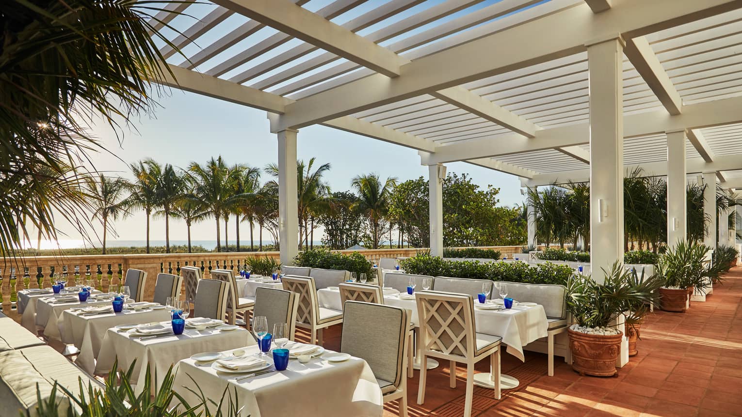 Large restaurant patio with formal dining tables under white pergola. Palm trees and beach in background