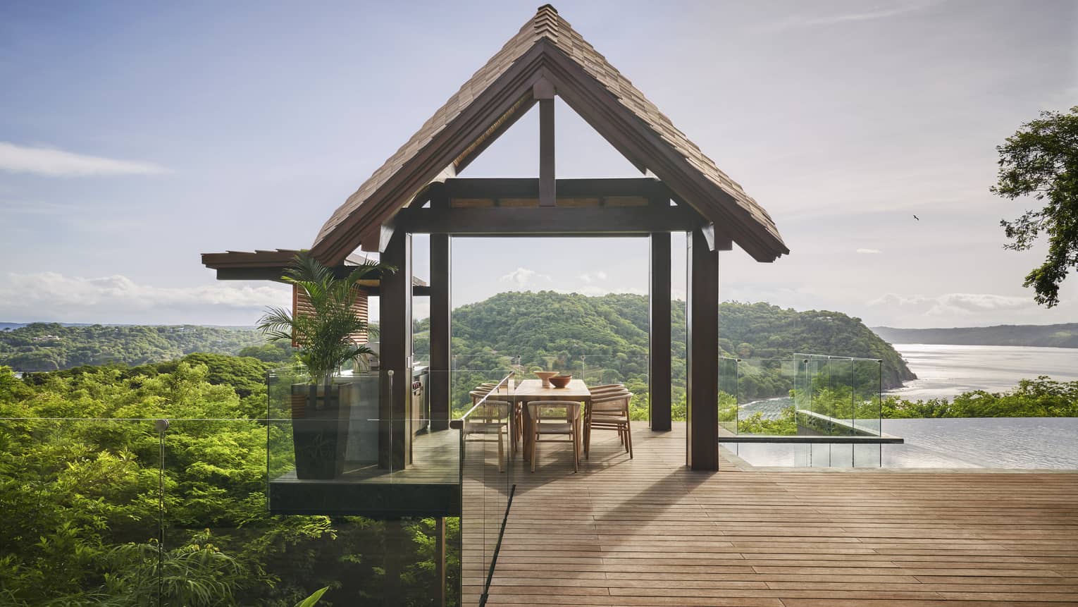 Dining table sits under an open-air wooden pavillon, next to a pool and overlooking the sea